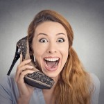 Headshot young happy woman looking excited, holding an high heeled shoe in her hand as a phone isolated on grey wall background. human face expression emotion feelings